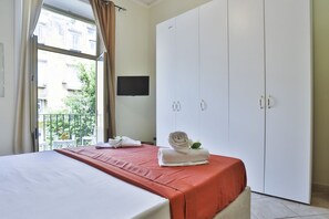 Ein Hotelzimmer mit Bett, großem Kleiderschrank, Fernseher an der Wand und einem Balkon mit Blick auf Gebäude und Grünflächen.