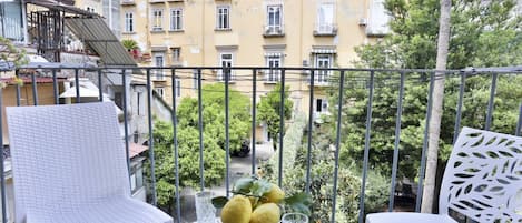 A balcony with a white chair, a table with a fruit bowl and glasses, and a view of a yellow building with multiple windows.