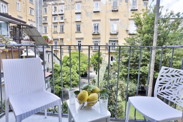 Ein Balkon mit einem weißen Stuhl, einem Tisch mit Obstschale und Gläsern sowie Blick auf ein gelbes Gebäude mit zahlreichen Fenstern.