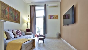 A living room with a sofa, a coffee table, a TV mounted on the wall, and a window with curtains. - Bright and welcoming apartment for three people, located in a historic building on one of Naples' most iconic streets. The two balconies overlooking the internal courtyard ensure tranquility and privacy despite its central location. It's perfect for expl (Napoli)