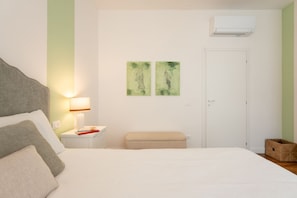 Ein Hotelzimmer mit Bett, Nachttisch, Lampe, zwei grünen Gemälden, einer weißen Tür und einer Klimaanlage.