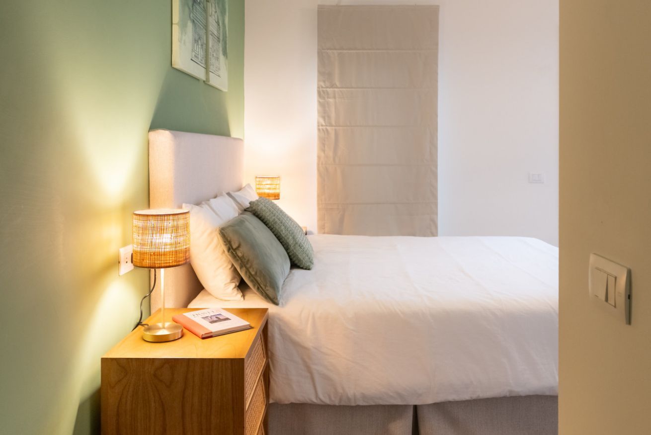 Un letto rifatto con cuscini, una lampada da comodino, un libro e una presa elettrica su un comodino in legno.