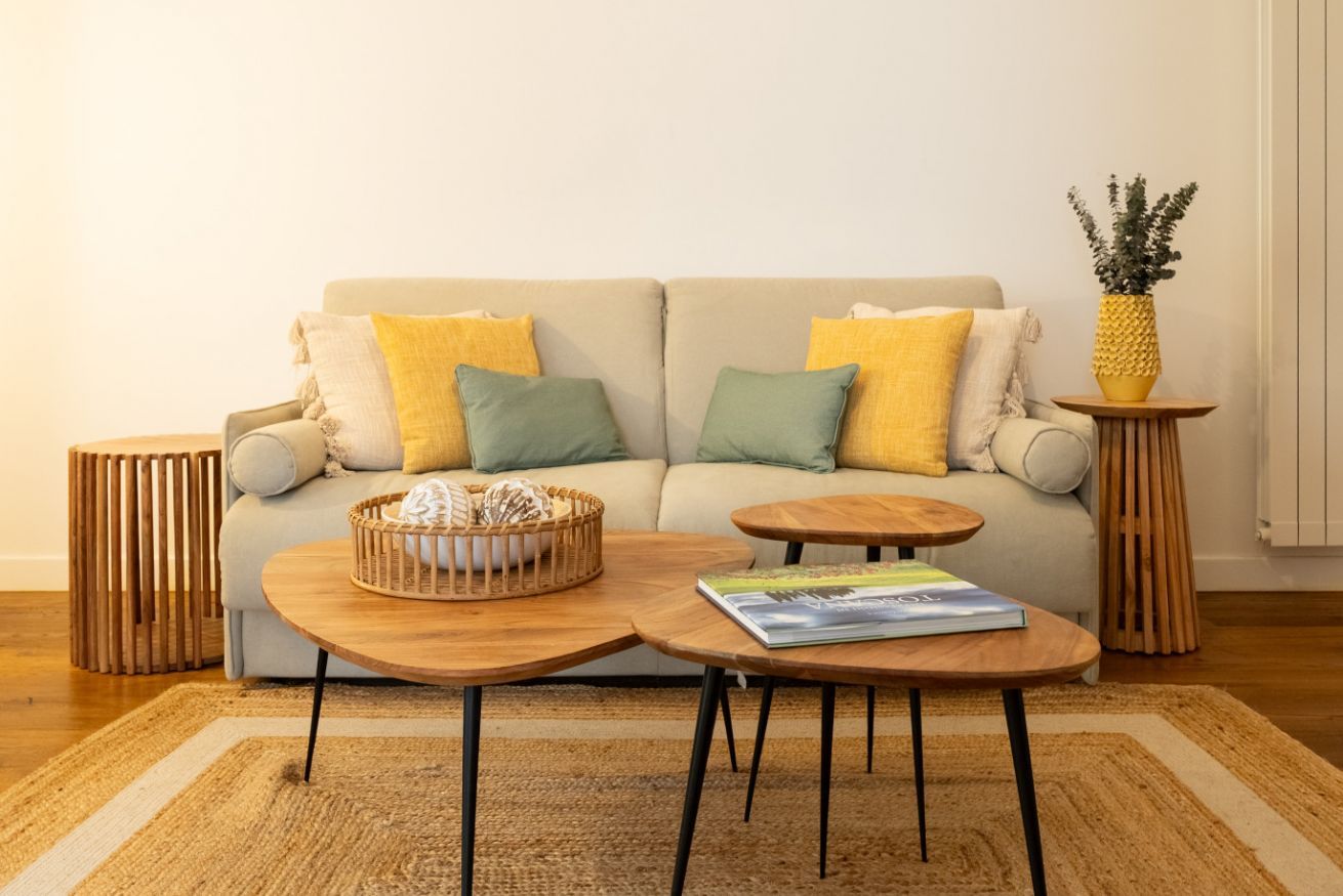 Een woonkamer met een bank, twee ronde houten tafels, een geweven mand en een klein houten bijzettafeltje met een plant.
