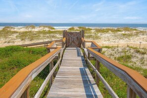 Ein hölzerner Steg führt zu einem Strand mit Dünen und dem Meer im Hintergrund.