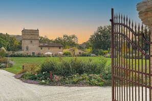 Un edificio in pietra con una torre, circondato da un giardino ricco di piante e fiori, e un cancello in metallo.