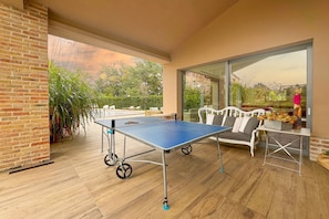 Una mesa de ping pong en una habitación soleada con pared de ladrillo, ventanas amplias y un sofá blanco.