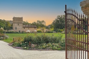 Ein Steinhaus mit einem Turm, umgeben von einem Garten mit verschiedenen Pflanzen und Blumen, und einem Metallzaun.