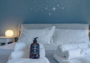A neatly made bed with white linens, a bottle of shampoo, and rolled towels on a blue wall with star decals.