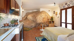 Cuina amb paret de pedra, llit amb estovatge blanc i zona de menjador amb taula i cadires.