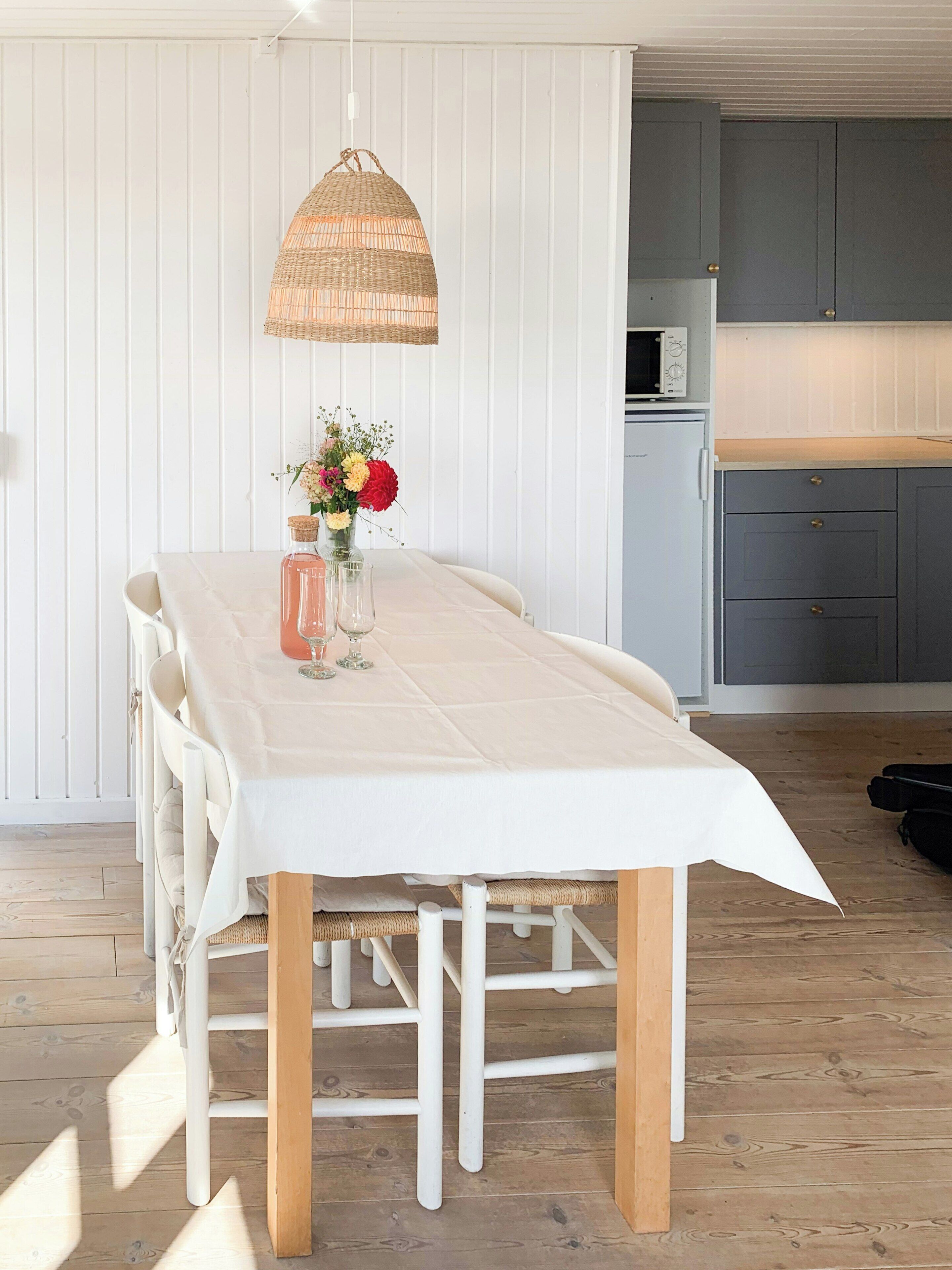 A dining table with a white tablecloth, a wicker hanging light, and a vase with flowers.