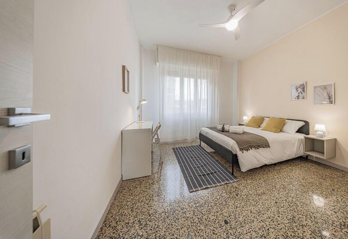 Ein Schlafzimmer mit Bett, Nachttischen, Schreibtisch, Stuhl, einem Fenster mit Vorhängen und einem Deckenventilator.