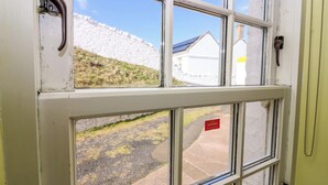 A window with a view of a white building, a grassy hill, and a paved area. - Lightkeepers Fold (Eyemouth)
