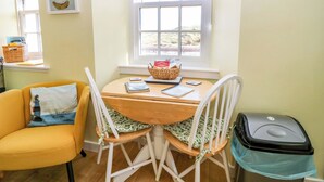 A cozy room with a wooden table, two white chairs, a yellow armchair, a window with a view, a clock, and a basket of bread on the windowsill. - Lightkeepers Fold (Eyemouth)