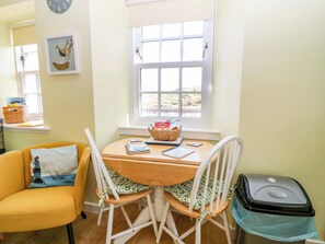 Une pièce chaleureuse avec une table en bois, deux chaises blanches, un fauteuil jaune, une fenêtre donnant sur un beau paysage, une horloge et un panier de pain posé sur le rebord de la fenêtre.