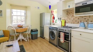 A compact kitchen with a washing machine, oven, and a microwave. There's a dining area with a table and chairs, and a window providing natural light. - Lightkeepers Fold (Eyemouth)