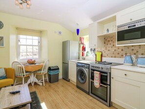 A compact kitchen with a washing machine, oven, and a microwave. There's a dining area with a table and chairs, and a window providing natural light. - Lightkeepers Fold (Eyemouth)