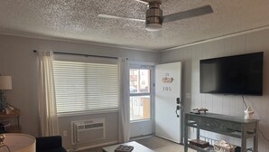 Ein Zimmer mit einer Couch, einem Couchtisch, einem Fernseher, einer Tür mit einer Nummer und einem Fenster mit Jalousien.