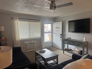 Ein Zimmer mit einer Couch, einem Couchtisch, einem Fernseher, einer Tür mit einer Nummer und einem Fenster mit Jalousien.