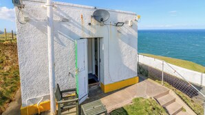 A small, white building with a door, a bench, and a view of the ocean. - Lightkeepers Flaghouse (Eyemouth)