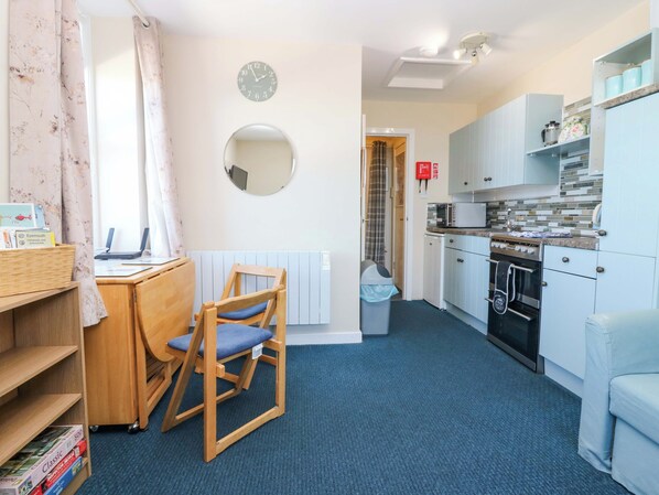 A compact kitchen with a microwave, stove, and cabinets, a wooden chair, a desk with books, a clock, and a mirror on the wall. - Lightkeepers Flaghouse (Eyemouth)