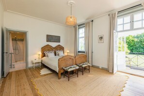 Une chambre avec un plancher en bois, un lit avec une tĂȘte de lit, deux tables de chevet avec des lampes, un luminaire suspendu et une grande fenĂȘtre avec des rideaux.