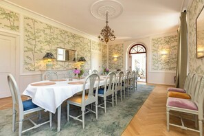 A dining room with a long table, chairs, a chandelier, and wallpaper with a leafy pattern.