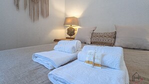 A neatly made bed with rolled white towels, a patterned pillow, a wall-mounted macrame piece, and a table lamp.
