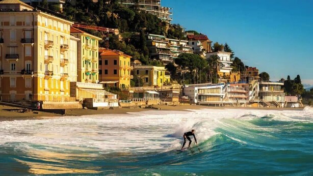 A coastal town with buildings along the shore, a person surfing, and waves crashing onto the beach.