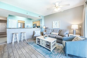 Ein Wohnzimmer mit einer Couch, einem Couchtisch und Hocker. Es gibt eine Küchenzeile mit Schränken und einer Mikrowelle. Ein Deckenventilator ist sichtbar.