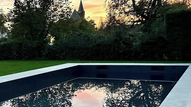 Ein Swimmingpool mit deutlicher Spiegelung von Bäumen und einem Kirchturm im Hintergrund.