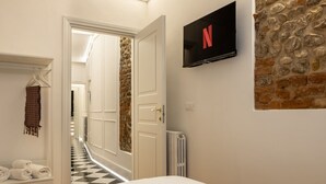 A hotel room with a bed, a TV mounted on the wall, a stone wall, a hallway with a checkered floor, and a door leading to another room. - A cozy and charming home featuring beautiful stone details, bright interiors, and a modern, relaxing atmosphere. Conveniently located in a central area, Atipico Apartment is perfect for guests who want to experience Florence with style and comfort.✔ Pe (Firenze)