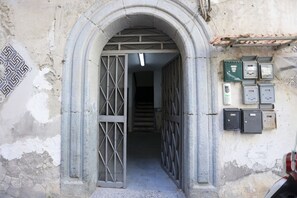 Une porte en arc surmontée de grilles métalliques, un escalier menant à l’intérieur et des boîtiers électriques fixés au mur.