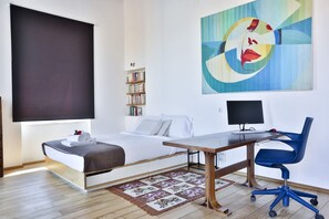 Ein modernes Hotelzimmer mit Bett, Schreibtisch, Stuhl, Bücherregal und einem großen abstrakten Gemälde.