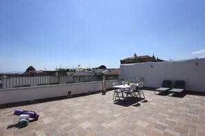 Eine Dachterrasse mit Tisch und Stühlen, ein überdachter Bereich mit Liegestühlen und Blick auf Gebäude in der Ferne.