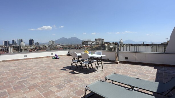Terraço no telhado com mesa e cadeiras, uma poltrona de descanso verde e vista panorâmica da cidade ao fundo.