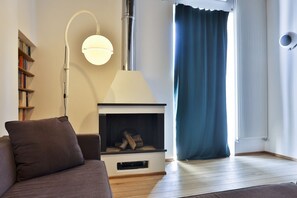 Ein Wohnzimmer mit Sofa, Kamin, Lampe, Bücherregal und einem fensterbehangenen Fenster.