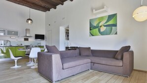 Uma sala moderna com sofá seccional, área de jantar com cadeiras brancas, cozinha com armários verdes e um quadro abstrato grande na parede.
