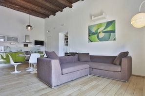 Ein modernes Wohnzimmer mit einem Ecksofa, einer Essecke mit weißen Stühlen, einer Küche mit grünen Schränken und einem großen abstrakten Gemälde an der Wand.
