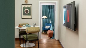 Une pièce comprenant une table à manger, des chaises, un tableau encadré, une lampe, une chaise bleue, un pouf rouge et un téléviseur fixé au mur.