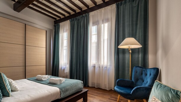 Une chambre à coucher avec un lit, un banc, une chaise, une lampe et une fenêtre avec des rideaux.