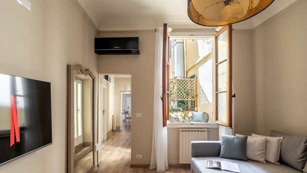 A modern living room with a grey sofa, a flat-screen TV, and a window with a view of the outside. - Augustus Florence Borgo Pinti 3 min Duomo (Firenze)