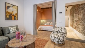 Ein modernes Wohnzimmer mit einer Couch, einem gemusterten Sessel, einem Couchtisch mit Dekoration und einem Bild an der Wand. Durch eine offene Tür ist ein Schlafzimmer sichtbar.