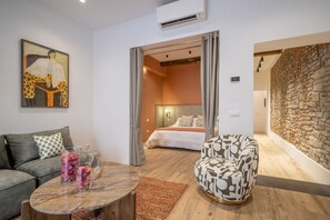 Ein modernes Wohnzimmer mit einer Couch, einem gemusterten Sessel, einem Couchtisch mit Dekoration und einem Bild an der Wand. Durch eine offene Tür ist ein Schlafzimmer sichtbar.