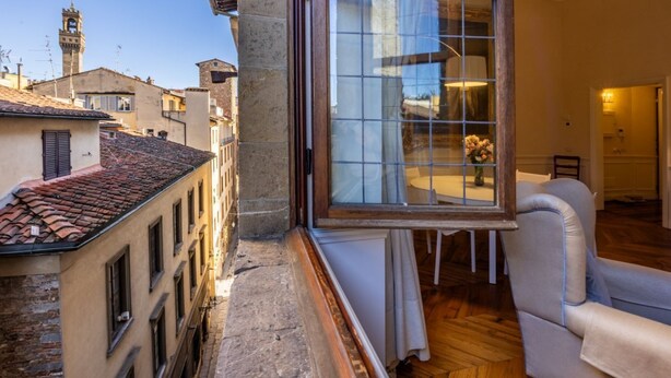 A room with a large window overlooking a cityscape, a chair, and a table with a vase of flowers. - The Loggia of Borgo SS Apostoli (Firenze)