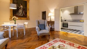 A room with a dining table, chairs, a sofa, a large framed painting, a chair with a cushion, a lamp, and a kitchen area with a checkered floor. - The Loggia of Borgo SS Apostoli (Firenze)