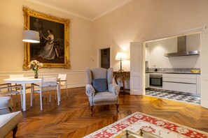 Ein Zimmer mit Esstisch, Stühlen, einer Couch, einem großen gerahmten Gemälde, einem Sessel mit Kissen, einer Lampe und einer Küchenzeile mit kariertem Boden.