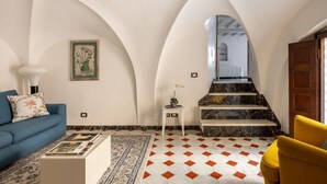 Ein Wohnzimmer mit einer Couch, einem Couchtisch, einem gemusterten Teppich, einer Treppe mit Marmorverkleidung und einem gerahmten Bild an der Wand.
