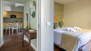 A hotel room with a bed, a desk, a chair, a small table, and a kitchen area visible through an open door. - Geometric Design Suite by Maison Florence (Florence)