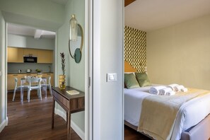 A hotel room with a bed, a desk, a chair, a small table, and a kitchen area visible through an open door. - Geometric Design Suite by Maison Florence (Florence)