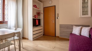 Una habitación con piso de madera, una cama con cubrecolcha púrpura, un televisor en un estante, una puerta de madera y una ventana con cortinas.